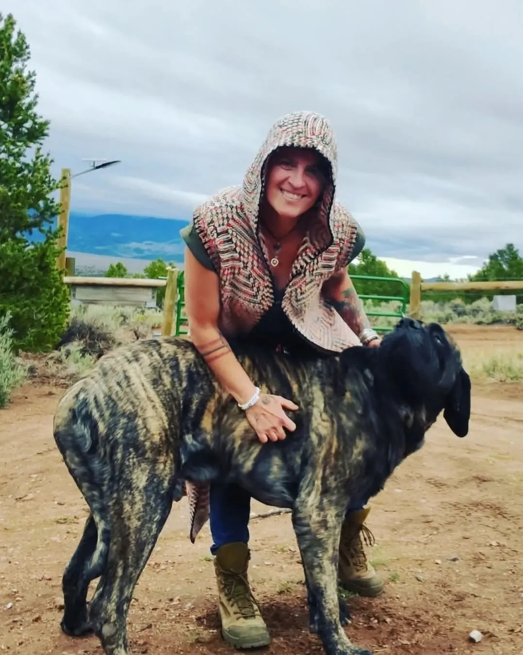 Cheryl smiling in a hooded knit vest, leaning over a large brindle dog in a ranch yard with mountains behind